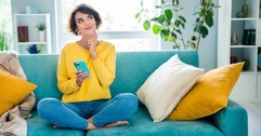 A girl is thinking while sitting on the couch in the living room and holding her phone.