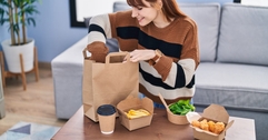 A woman sits at a coffee table and unpacks a food delivery order. She looks happy.