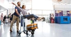 Woman sitting on luggage on a cart being pushed by a man through an airport