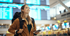 A woman wears a backpack and holds her phone in the middle of an airport.