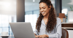 A woman smiles while typing on her laptop in an office setting.