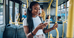 A woman wears headphone and smiles while using her smartphone on a bus.