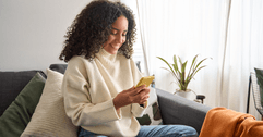 A woman in a creme colored sweater uses her phone while sitting on a gray couch.