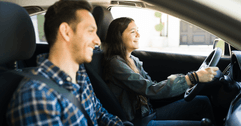 A woman smiles while driving her car, as a man smiles in her passenger seat.