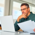 Man in his 40's looking at paperwork and computer thinking