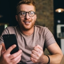 A man wearing glasses sits in front of a laptop and holding a smartphone. 