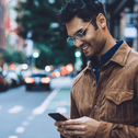 A man wearing glasses stands on the sidewalk in a busy city street while smiling and staring down at his phone.