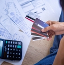 A person holding four credit cards sits in front of a pile of bills and a calculator. Their hand is on their face as if they&#x27;re thinking.