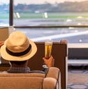 A man sits in a chair at the airport holding a beer and touching the brim of his hat. 