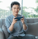 A man wearing headphones sits on a couch and plays mobile games on his phone. 