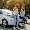 Two girls leaning on white electric car waiting while it&#x27;s charging at station green trees and sun 