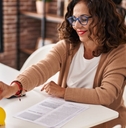 middle age woman sitting at table with papers putting pennie into yellow piggy bank