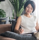 asian woman sitting on couch at home writing on notepad with pen while using laptop
