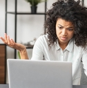 african american woman sitting in front of laptop at table raising hands in confusion