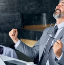 senior businessman sitting at table in front of laptop cheering in happiness while looking up