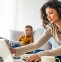A mom works from her laptop while sitting on the couch next to a young child. 