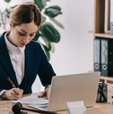 female lawyer wearing suit sitting in office writing on papers while using laptop