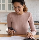 happy woman sorting out budget in kitchen while holding smartphone