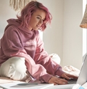 A young woman sits cross-legged on a bed and looks at a laptop screen in front of her. 