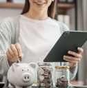 A young woman is holding an iPad with one hand while inserting a coin into her piggy bank with another hand.