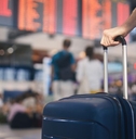A mostly unseen person stands in front of a flight board in an airport, with one hand holding onto the handle of their suitcase.