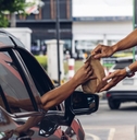 A car at a drive-thru window. A hand outstretched from the car takes a paper bag of food from a drive-thru worker who is handing it over.