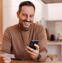 A man in a brown turtleneck smiles while using his phone and holding a credit card in his other hand.