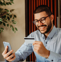 A young man wearing glasses smiles while holding a credit card and looking at his phone.