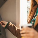 Woman wearing leather jacket and holding a drink using her bank card in an ATM