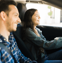 A woman smiles while driving her car, as a man smiles in her passenger seat.