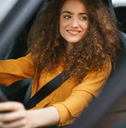A woman with curly hair and an orange shirt grips the steering wheel and smiles.