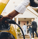 A woman charges a yellow electric vehicle while her family walks into their home in the background.