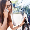 A woman calls someone on her iPhone while touching an indent near the rear wheel of her vehicle.