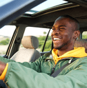 A man in a green jacket smiles and laughs as he grips the steering wheel.