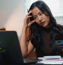 A woman holds a credit card and grips her head in frustration as she looks at bills on her computer. 