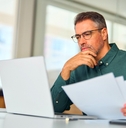 Man in his 40's looking at paperwork and computer thinking