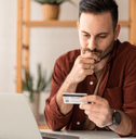 A man appears worried as he holds his chin and looks down at his credit card while sitting in his home office.