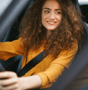 A woman with curly hair and an orange shirt grips the steering wheel and smiles.