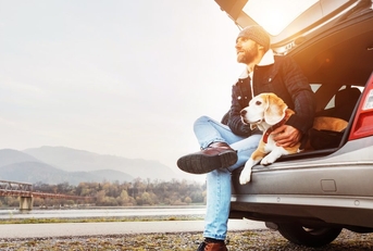 A man and a dog sit on the edge of the cargo area in a hatchback car. 