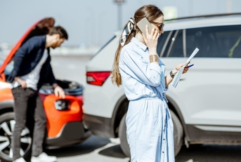 A woman makes a phone call while a man inspects a fender bender car accident behind her.