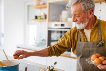 happy senior man cracking eggs and cooking pasta in kitchen