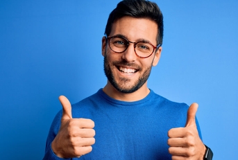 young man in blue t shirt wearing glasses doing thumbs up in front of blue background