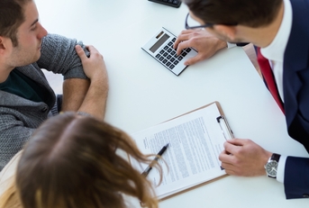 Couple looking at paper forms and calculator on the table being used by man in suit on the other side of the table