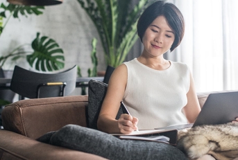 asian woman sitting on couch at home writing on notepad with pen while using laptop