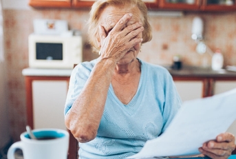 senior woman covering her face in stress while looking at utility bills in kitchen