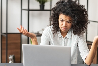 african american woman sitting in front of laptop at table raising hands in confusion