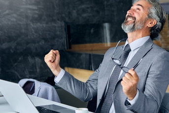 senior businessman sitting at table in front of laptop cheering in happiness while looking up