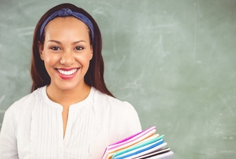 A woman teacher stands in front of a chalkboard holding a pile of colorful folders. She&#x27;s smiling.