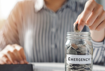woman putting pennies in glass jar with emergency label on it while using calculator to finance mortgage bills