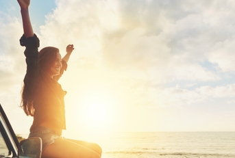 woman on road trip sitting on car bonnet at beachside raising hands in the air